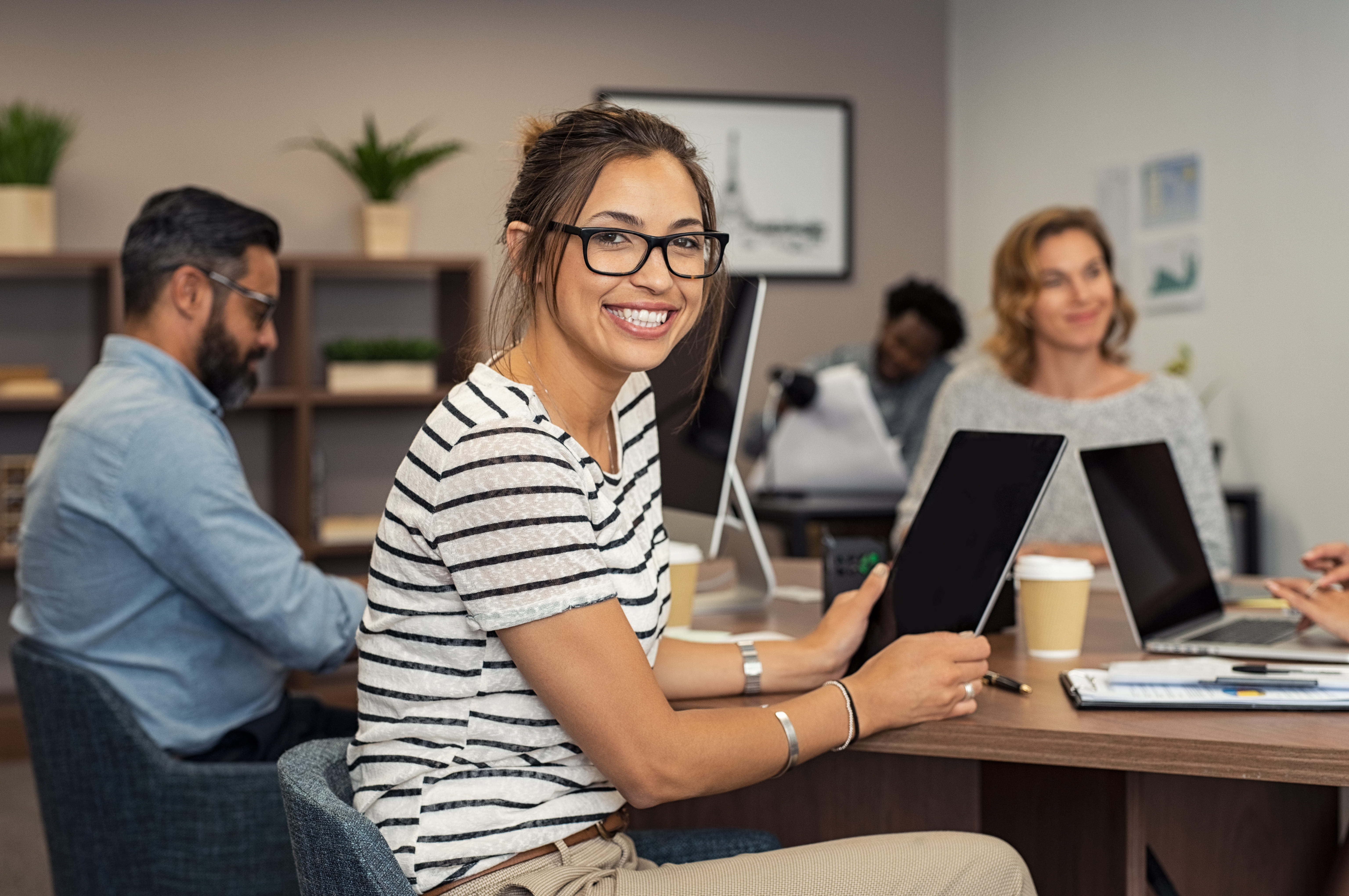 Smiling woman with glasses holding a tablet in a collaborative office environment with colleagues.