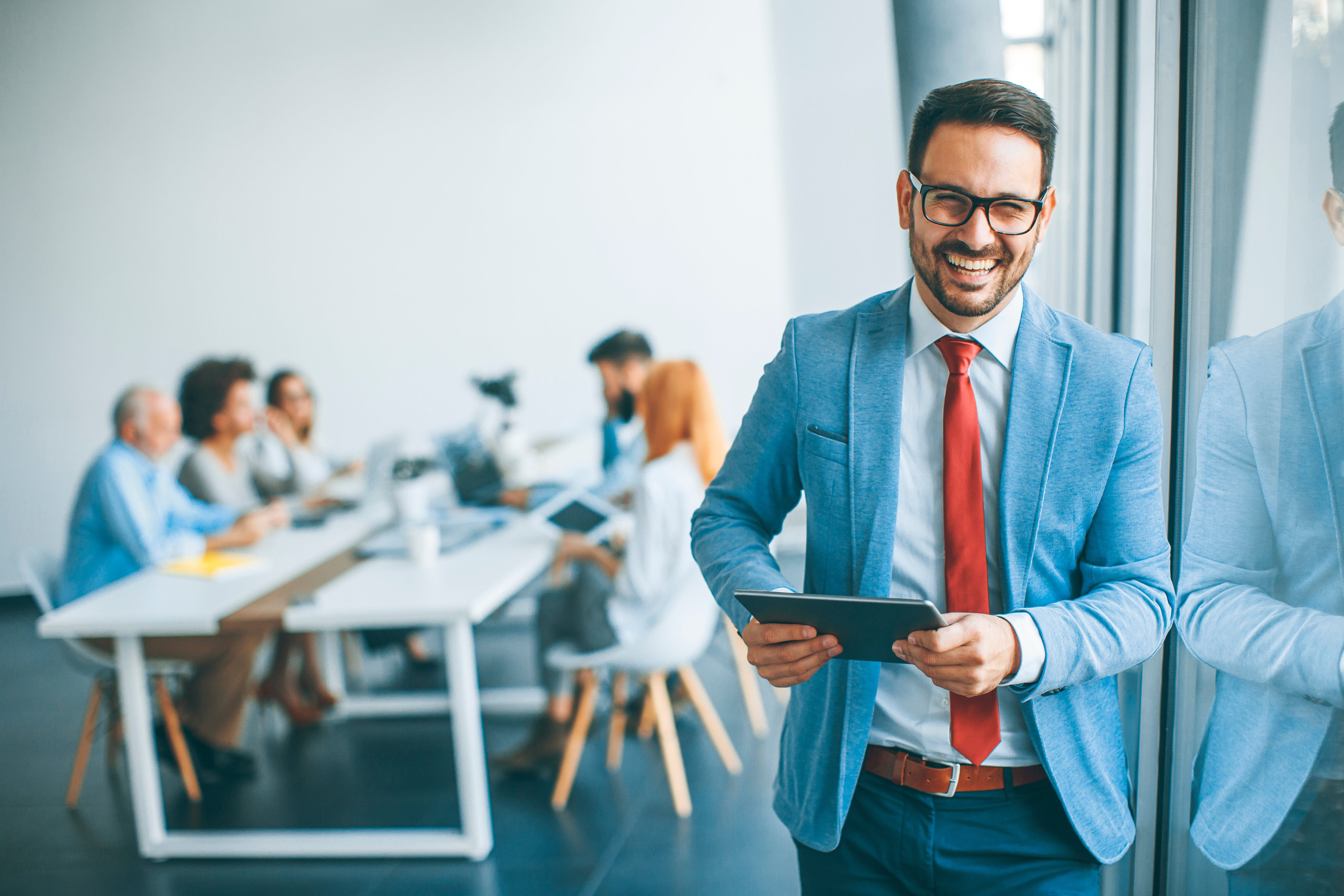 Smiling businessman holding a tablet in the foreground with a team meeting in progress in the background.