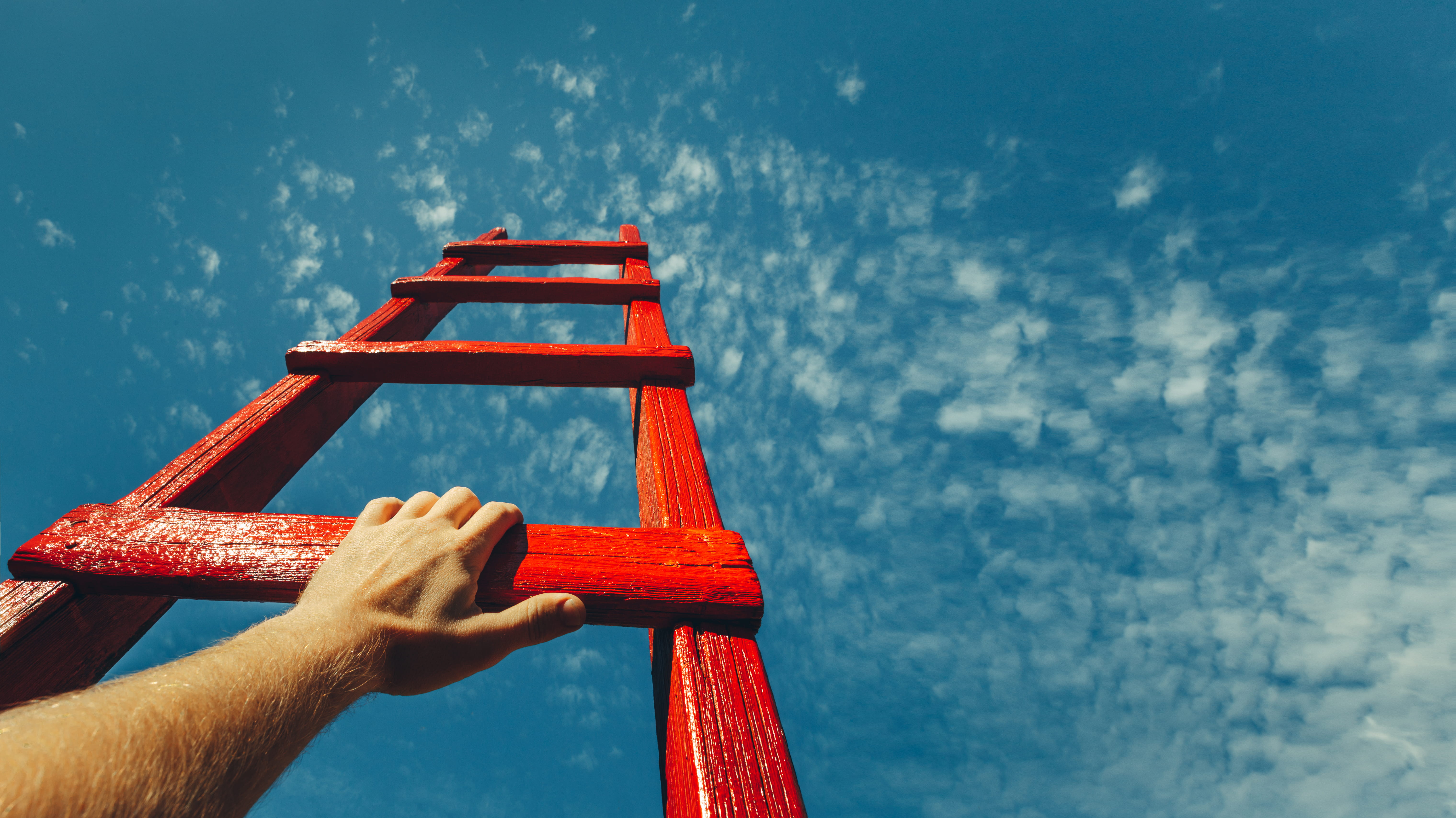 Hand holding a red ladder rung, extending upwards towards a bright blue sky with scattered clouds