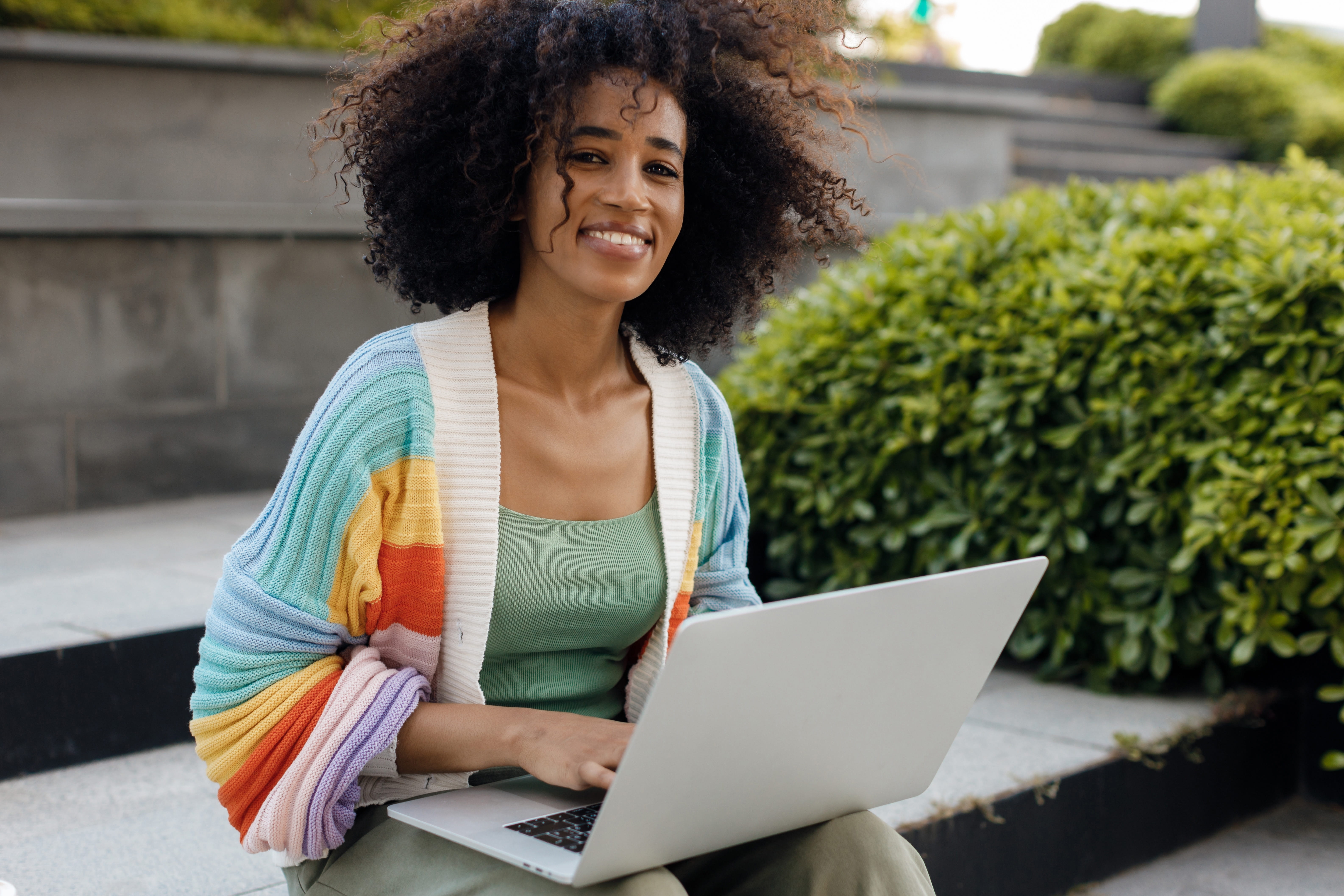 A smiling person working on a laptop while sitting outdoors in a landscaped area.