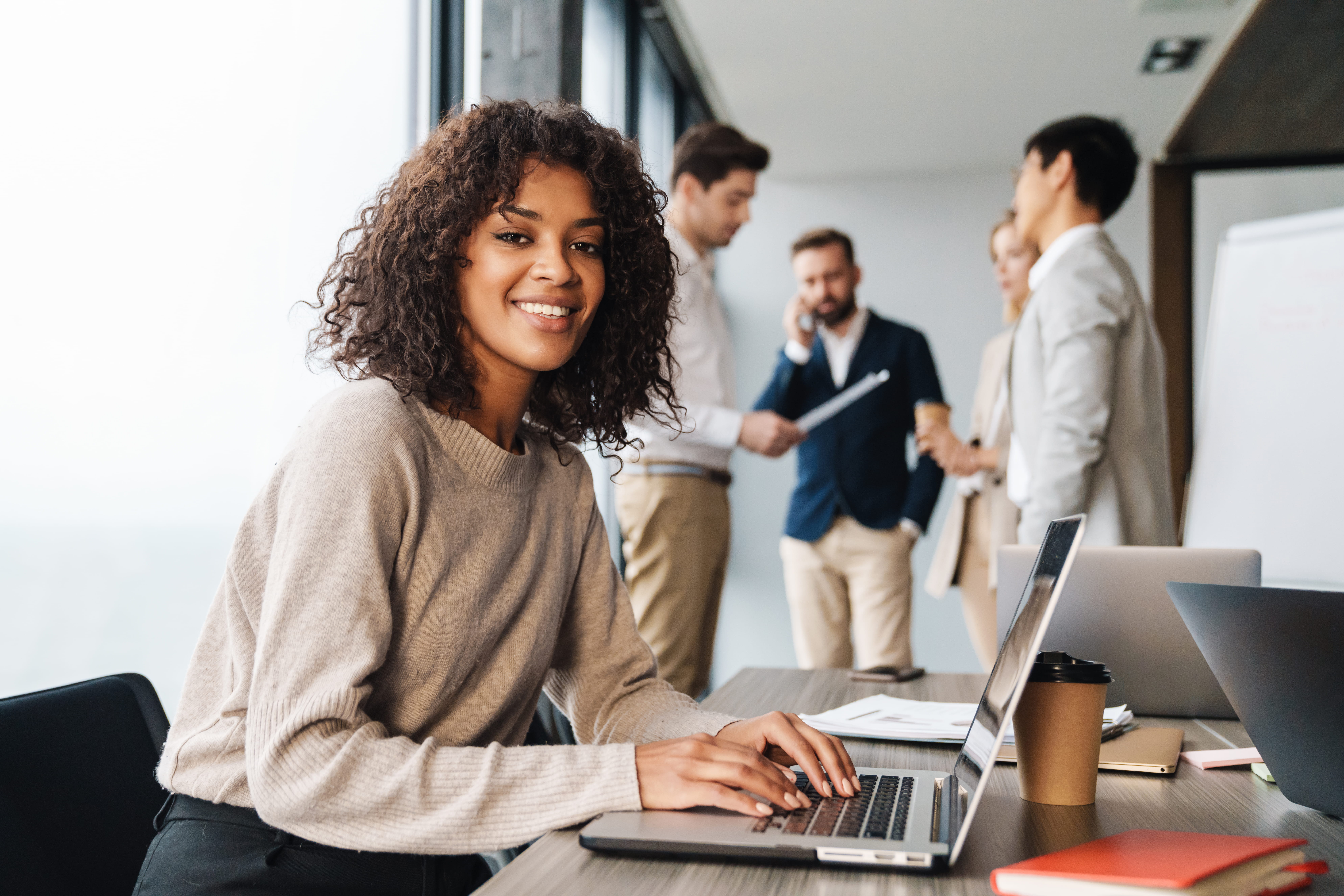 Smiling person working on a laptop seated at desk with coworkers in the background. Smiling person working on a laptop seated at desk with coworkers in the background.