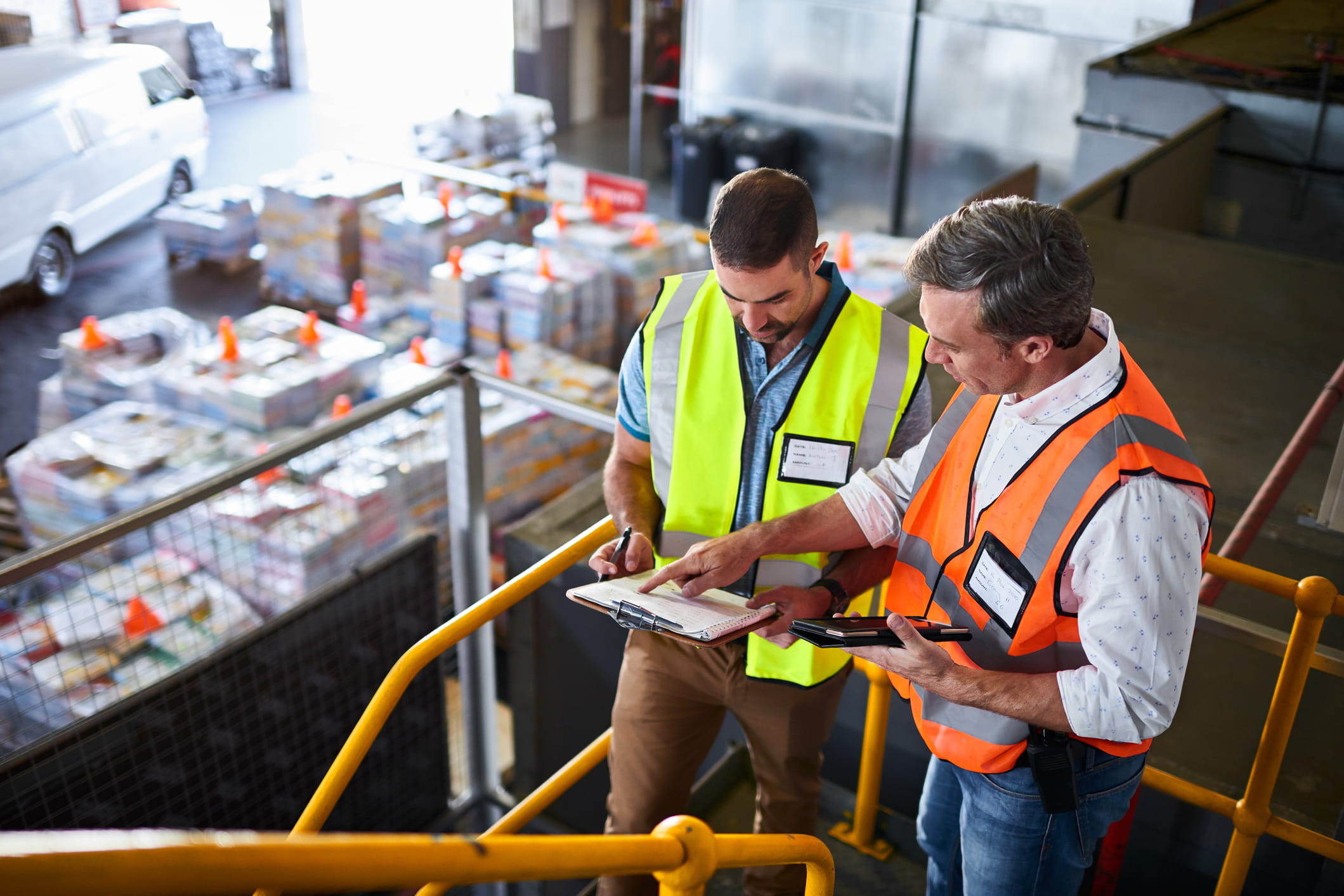 Two workers wearing high-visibility vests are standing on a staircase in a warehouse. 