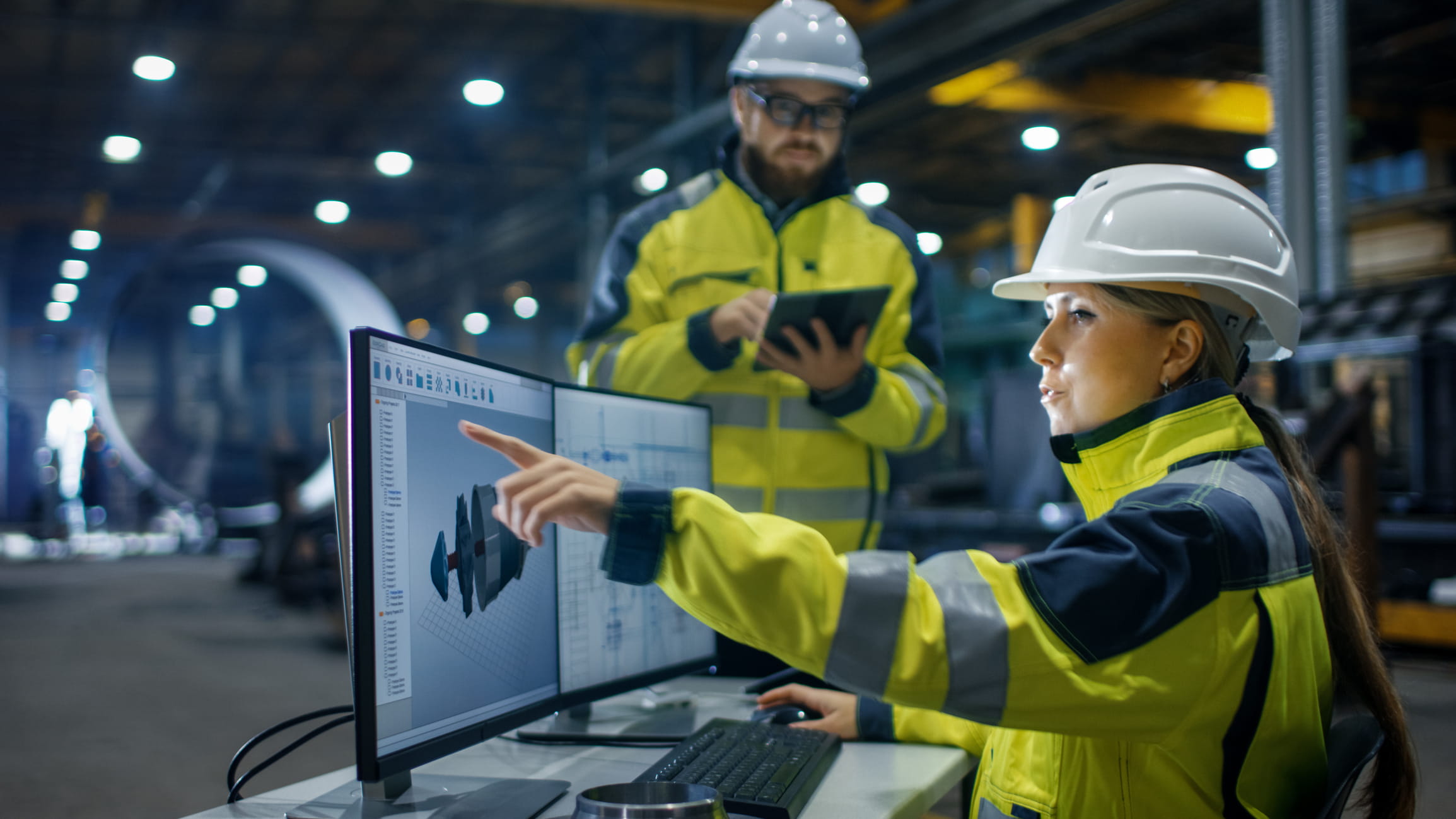 Two individuals wearing high-visibility jackets and white hard hats are in an industrial setting looking at a computer screen.