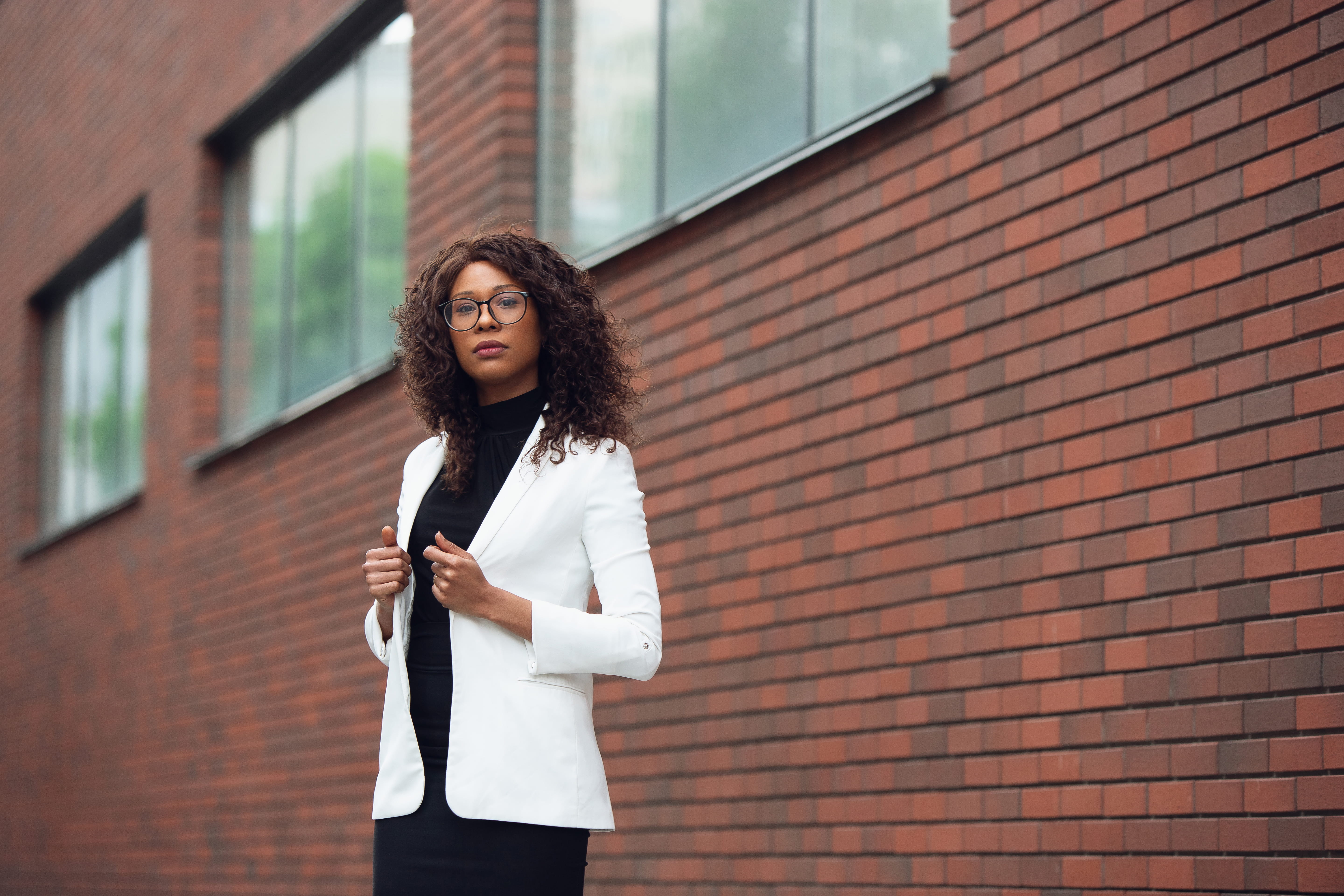 Confident woman in a white blazer standing against a brick wall, looking at the camera.