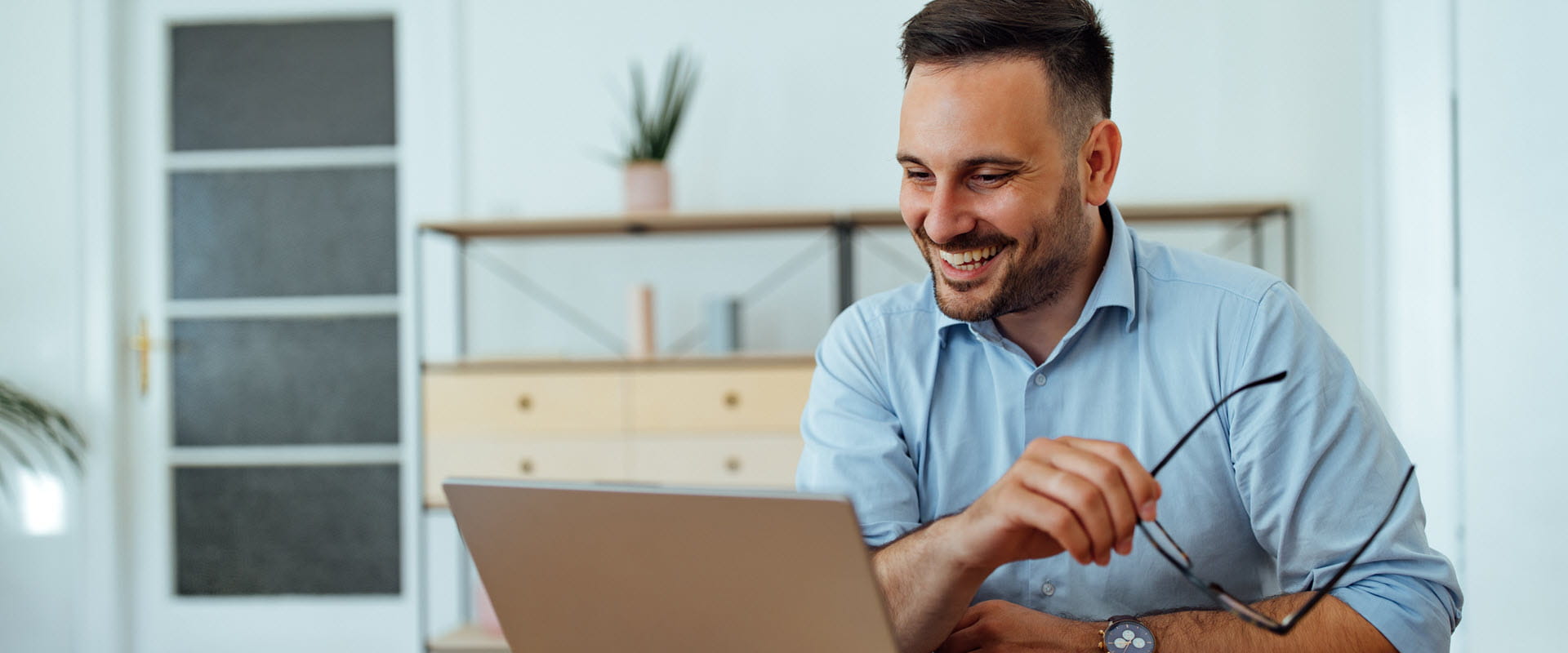 A smiling individual sitting at a table, engaging in a virtual meeting on a laptop while holding glasses in one hand. A smiling individual sitting at a table, engaging in a virtual meeting on a laptop while holding glasses in one hand.