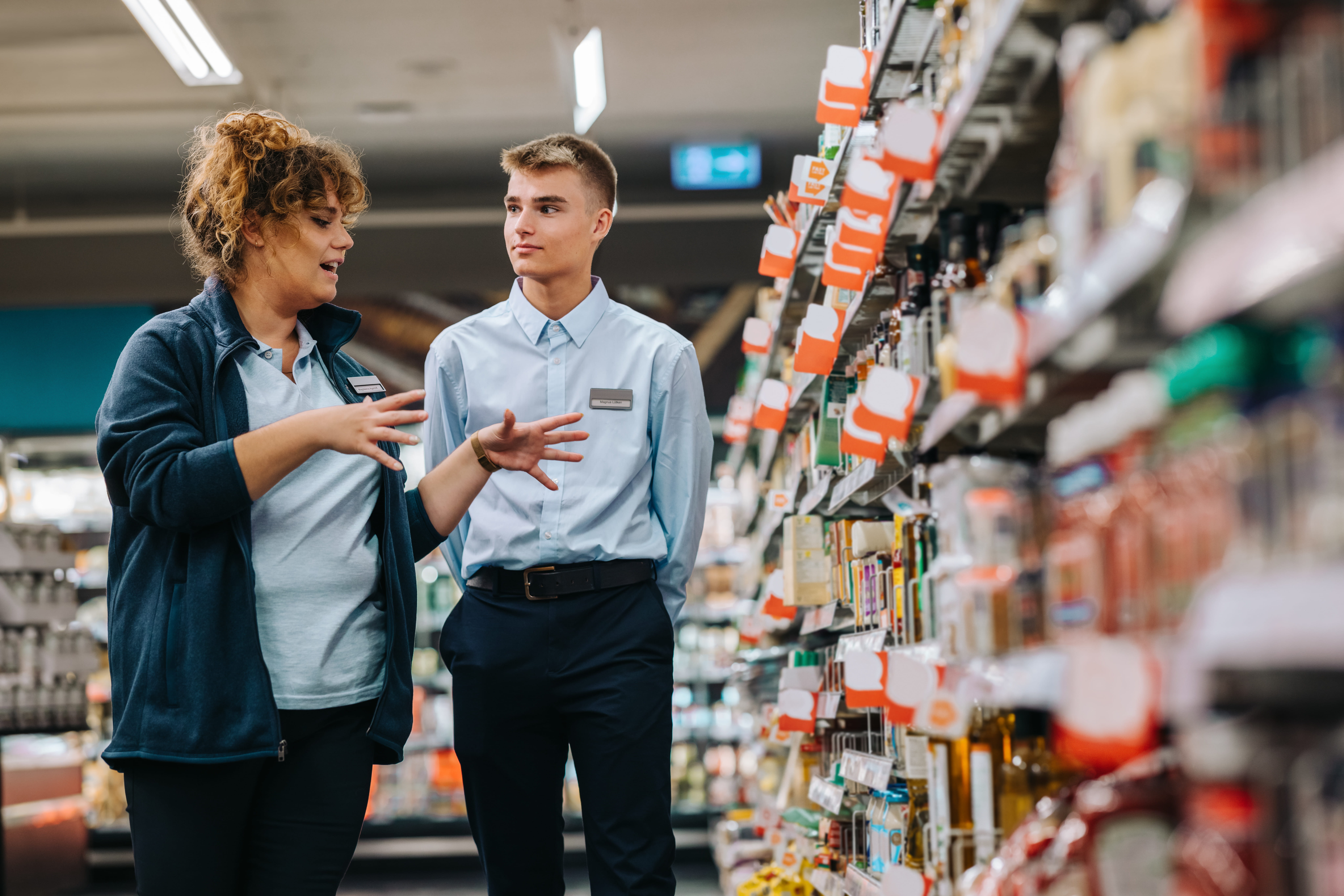 Retail store merchandisers discuss item sales and stock deliveries in an aisle full of neatly displayed products.