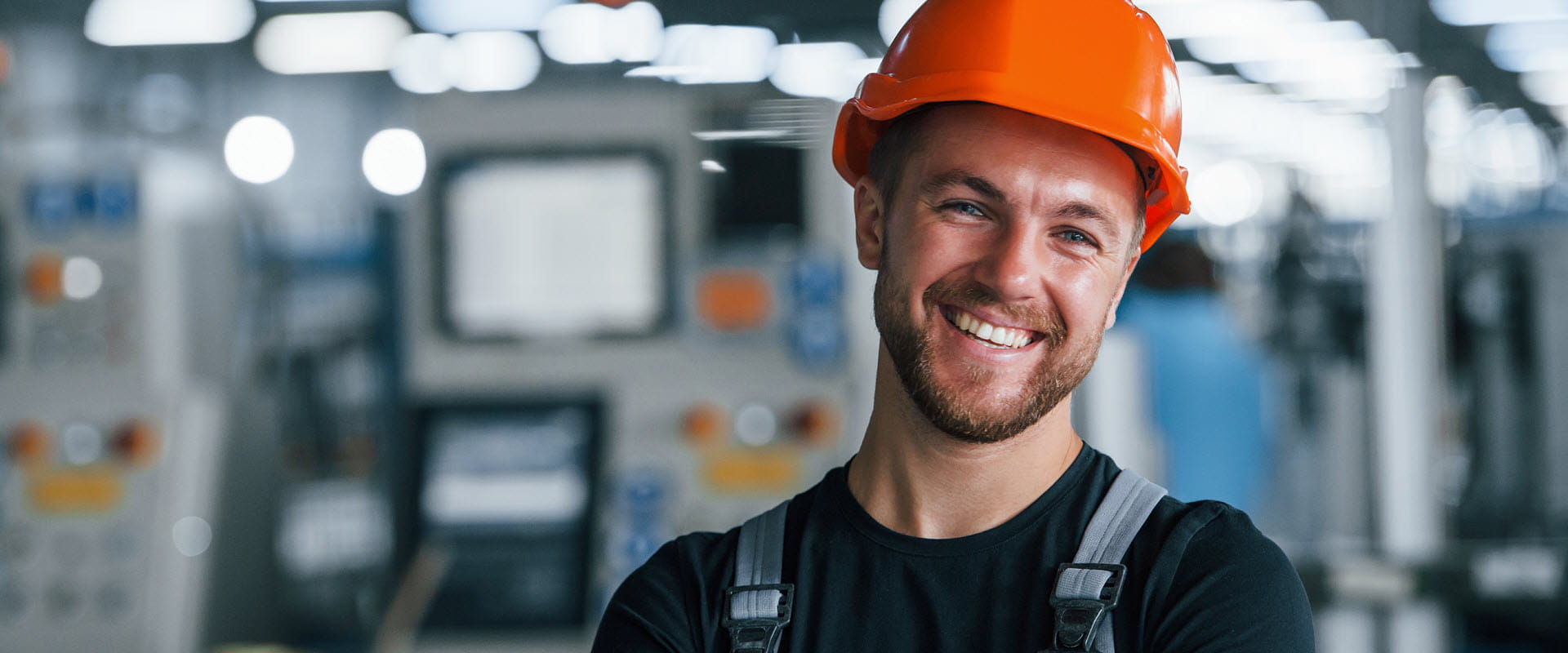 Smiling worker in an industrial setting, wearing an orange hard hat and black work clothes.