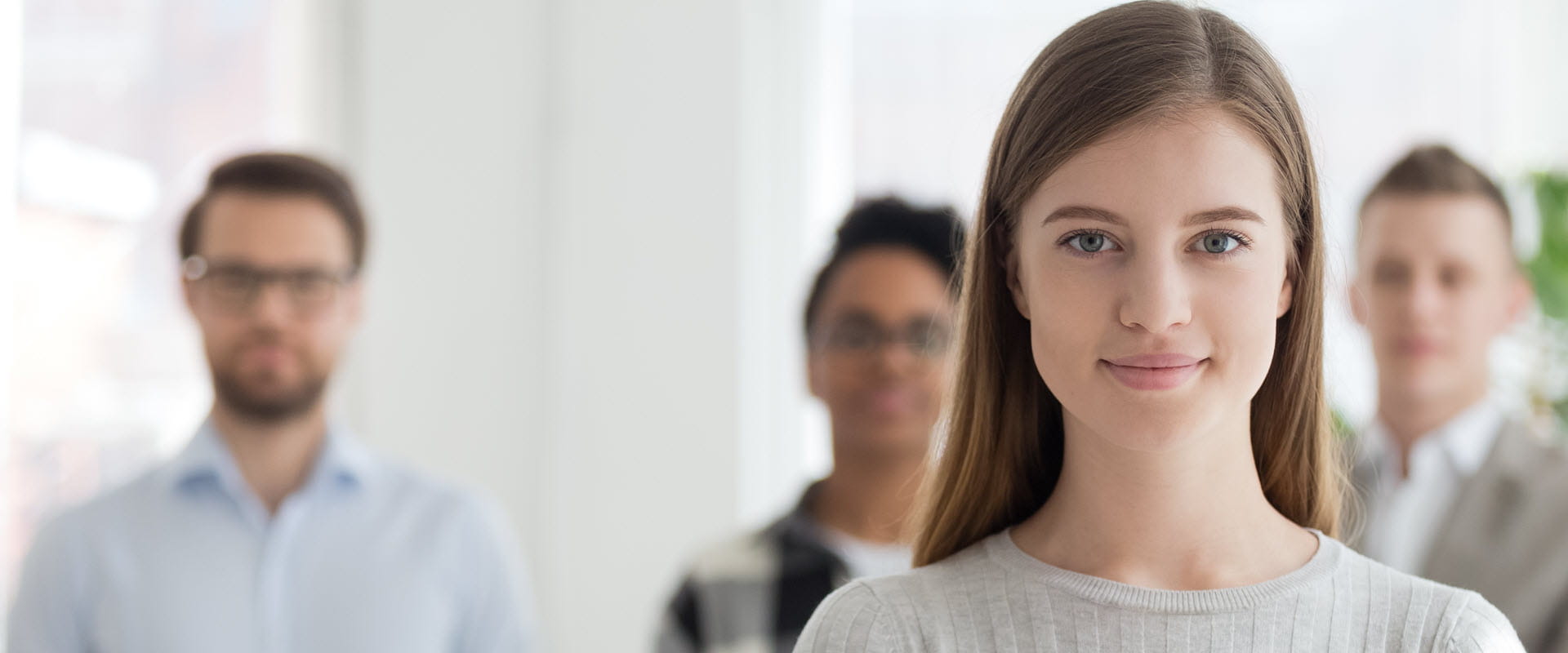 A confident young professional standing in front of their team members, who are slightly blurred in the background. A confident young professional standing in front of their team members, who are slightly blurred in the background.